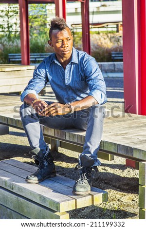 Modern Worker.  Wearing a blue shirt,  gray pants,  leather boot shoes, a young black guy with mohawk hair is sitting on a wooden structure, looking and thinking.