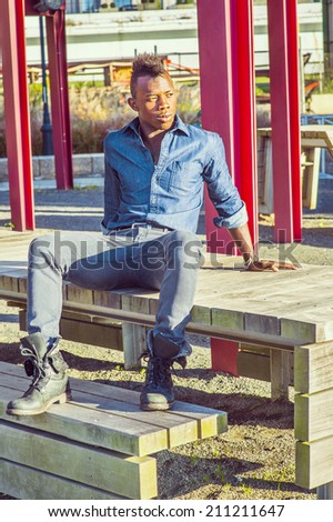 Young Worker Relaxing Outside.  Wearing a blue shirt,  gray pants,  leather boot shoes, a young black guy with mohawk hair is sitting back on a wooden structure, looking and thinking.