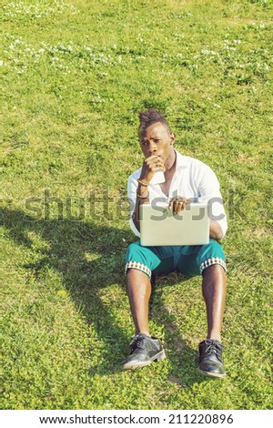 Dressing in a white shirt,  green pants,  black leather shoes,  holding a laptop computer and a mobile phone, a young black guy with mohawk hair is sitting on a green grasses, thinking.