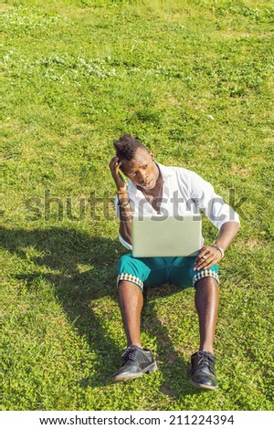 Dressing in a white shirt,  green pants,  black leather shoes,  holding a laptop computer and a mobile phone, scratching head, a young black guy with mohawk hair is sitting on green grasses, thinking.