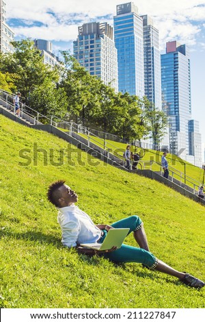 Man Working Outside. Dressing in a white shirt, green pants, black leather shoes,  a young black guy with mohawk hair is lying on a green hill in a big city, working on a laptop computer.