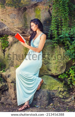 Pretty Lady Reading Outside. Dressing in a light blue long dress, a young college student with long curly hair is standing by rocky wall with long green leaves, concentrateingly reading a red book.