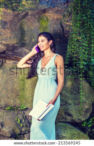 Modern Life. Dressing in a light blue dress, a young pretty woman with long curly hair is standing by rocky wall with long green leaves, holding a book, smiling, taking a phone call. Leisure Time,