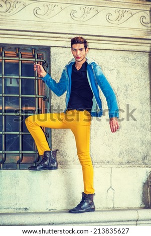 Man Casual Fashion. Dressing in blue jacket with hood, black underwear, yellow pants, leather boot shoes, a young handsome guy is standing by a window, foot on the frame, confidently looking at you.