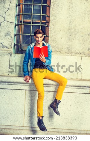 Man Reading Outside. Dressing in a blue jacket with hood, black under wear, yellow pants, leather boot shoes, holding a red book, a young handsome guy is sitting by a window, looking down, reading