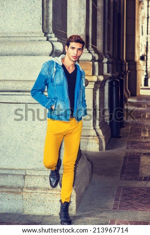 Man Relaxing Outside. Dressing in a blue jacket with hood, black underwear, yellow pants, leather boot shoes, hands in pockets, a young guy is standing by a column, looking at you. Street Fashion.