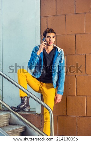 Worker Calling Outside. Wearing a blue jacket with hood, yellow pants, leather boot shoes, a young handsome guy is standing by railing in the front of office door way, talking on a mobile phone.