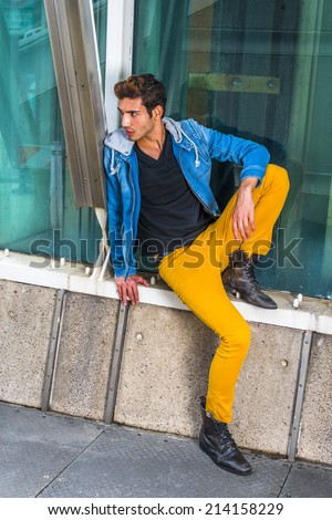 Man Waiting for You. Dressing a blue jacket with hood, black under wear, yellow pants, leather boot shoes, a young handsome guy is sitting by a metal structure, against a glass wall, looking around.