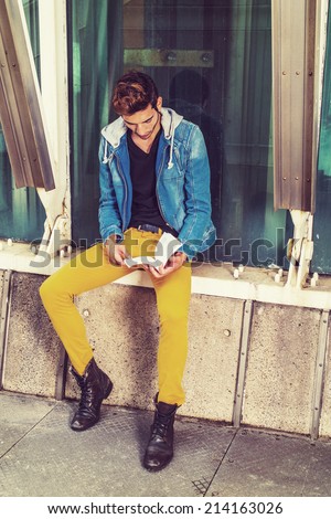 Man Reading Outside. Dressing in a blue jacket with hood, black under wear, yellow pants, leather boot shoes, a young handsome guy is sitting by a metal structure, against glass wall, reading a book.