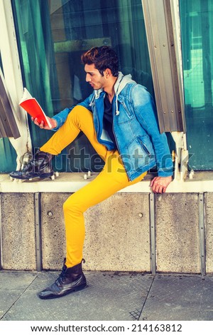 Man Reading Outside. Dressing in a blue jacket with hood, black under wear, yellow pants, leather boot shoes, a young guy is sitting by a metal structure, against a glass wall, reading a red book.