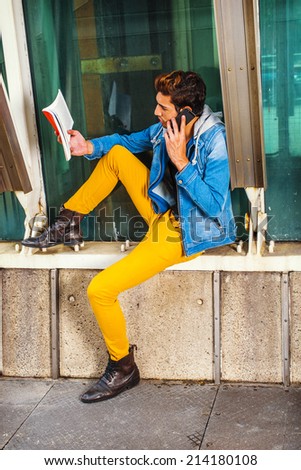 Man Busy. Dressing in a blue jacket with hood, yellow pants, leather boot shoes, a young handsome guy is sitting by a metal structure, reading a book and making a phone call in the same time.
