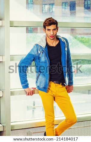 Portrait of College Student. Dressing in a blue jacket with hood, black under wear, yellow pants, a young handsome guy is standing against a glass wall, confidently looking at you.