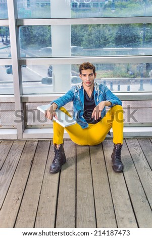 College Student. Dressing in a blue jacket with hood, yellow pants, black under wear, leather boot shoes, holding a white book, a young guy is sitting on wooden floor against a glass wall, relaxing,