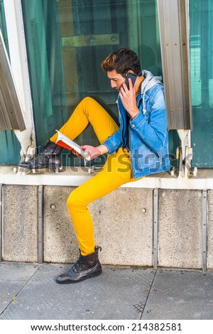 Busy Man. Dressing in a blue jacket with hood, yellow pants, leather boot shoes, a young handsome guy is sitting by a metal structure, reading a book and making a phone call in the same time.