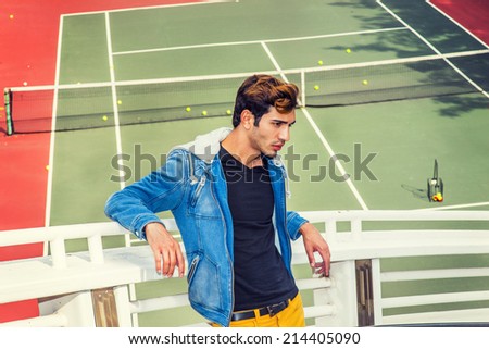 Man Lost in Thought. Dressing in blue jacket with hood, black under wear, yellow pants, a young handsome guy is standing by a tennis court, arms resting on railings, sad, thinking.