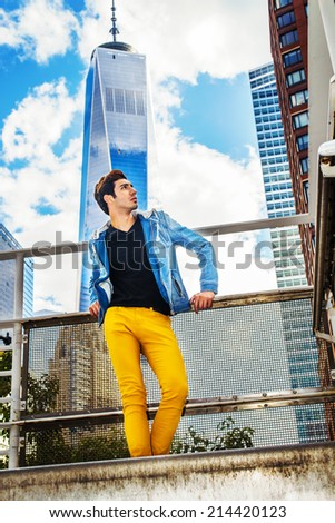 Looking for Success. Dressing in blue jacket with hood, black under wear, yellow pants, a young handsome guy is standing in the front of high tower building, confidently looking forward.
