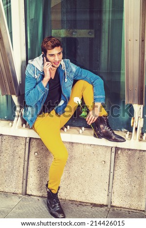 Man Waiting for You. Dressing in a blue jacket with hood, yellow pants, leather boot shoes, a young handsome guy is sitting by a metal structure, holding a white rose, talking on a mobile phone.