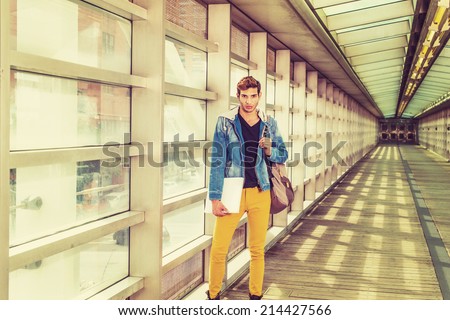 College Student on Campus. Dressing in a blue jacket with hood, black under wear, yellow pants, carrying a shoulder bag, holding a white book, a young guy is standing on walking way, looking at you.