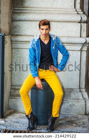 Man Casual Fashion. Dressing in a blue jacket with hood, black underwear, yellow pants, leather boot shoes, a young handsome guy is sitting on a metal stake on the corner, relaxing, thinking.
