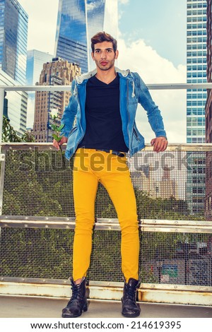 Man Waiting for You. Dressing in blue jacket with hood, yellow pants, leather boot shoes, holding a white rose, a young handsome guy is standing in the front of high buildings, looking forward.