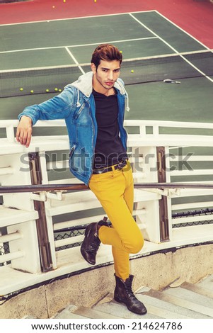 Man Casual Fashion. Dressing in blue jacket with hood, yellow pants, leather boot shoes, a young handsome guy is standing by a tennis court, arms resting on railings, relaxing, waiting, thinking.