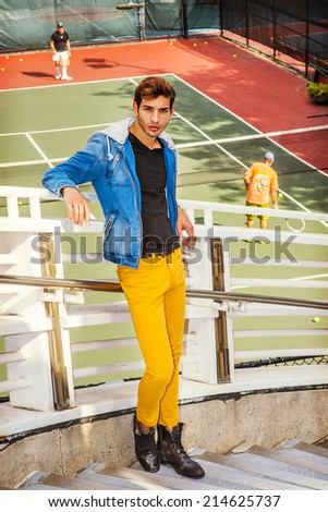 Man Thinking about You. Dressing in blue jacket with hood, yellow pants, leather boot shoes, a young handsome guy is standing by a tennis court, arms resting on railings, relaxing, waiting, thinking.