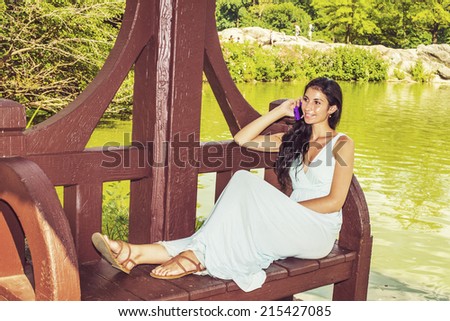 Dressing in a light blue, v neck, long dress, wearing  brown sandals, a young sexy woman with long curly hair is sitting on a red, long bench chair by a lake, smiling, talking on a mobile phone.