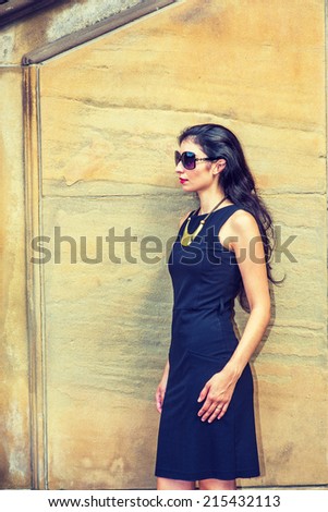 Business Lady. Dressing in black work dress, wearing sunglasses, a necklace with golden pendant, a young sexy businesswoman is standing outside, taking a break. Instagram effect.
