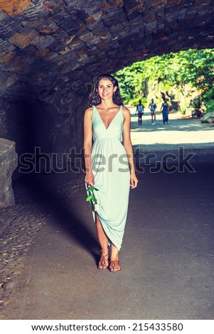 Holding a white rose, dressing in a light blue, v neck, long dress, wearing brown sandals, a young sexy woman is waking through a tunnel. Instagram effect.