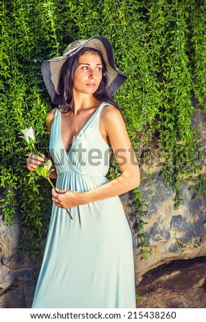 Lady Missing You. Dressing in light blue, v neck dress, wearing straw, wide brim hat, hands holding a white rose, a young sexy woman is standing by rocky wall with long green leaves, waiting for you.