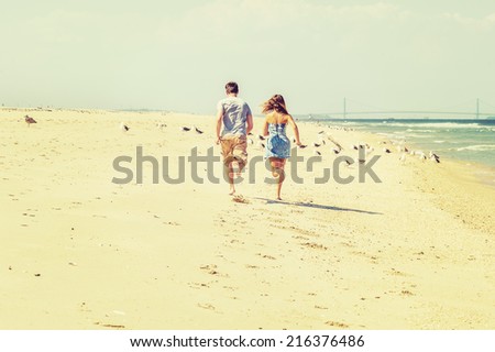 Young couple running on the sandy beach with barefoot in back view, guy wearing gray t shirt, yellow pants, girl dressing in strapless blue sun dress. Bridge, many birds in background.