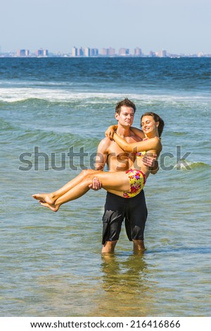 On the beach. Girl wearing a red, yellow patterned two piece bikini bathing suit, guy wearing a black bathing suit, guy holding girl in his arms, standing in water of ocean. Background is New York.