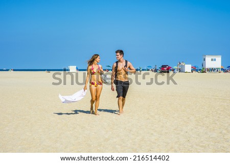 Go Back from Beach. Girl wearing a red, yellow patterned two piece bikini bathing suit, carrying a shoulder bag, a towel, guy wearing a black bathing suit, carrying a back bag, talking, walking.