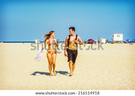 Go Back from Beach. Girl wearing a red, yellow patterned two piece bikini bathing suit, carrying a shoulder bag, a towel, guy wearing a black bathing suit, carrying a back bag, talking, walking.