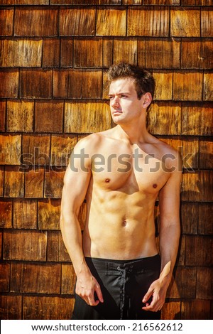 Portrait of Young Sexy Man. A well built young handsome muscular guy, half naked, wearing a bathing suit, is standing by a wooden wall on beach, narrowing eyes under sun shine, waiting for you.