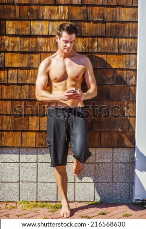 Texting Anywhere. A well built young handsome muscular guy, half naked, wearing a bathing suit, is standing by a wooden wall on beach, looking down, checking messages on his mobile phone.