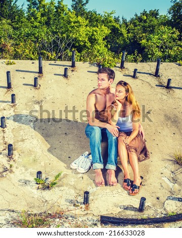 Story Teller. Guy wearing jeans, barefoot, half naked, girl dressing in skirt, tank top,  sunglasses on head, a young couple is sitting on the concrete slope with rusty metal rods, relaxing, thinking.