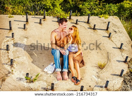Love Couple. Guy wearing jeans, barefoot, half naked, girl dressing in skirt, tank top,  sunglasses, a young couple is sitting on the concrete slope with rusty metal rods, looking each other, smile.