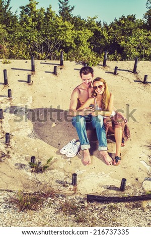 Love Couple. Guy wearing jeans, barefoot, half naked, girl dressing in skirt, tank top, sunglasses, a young couple is sitting on the concrete slope with metal rods, texting on mobile phone.