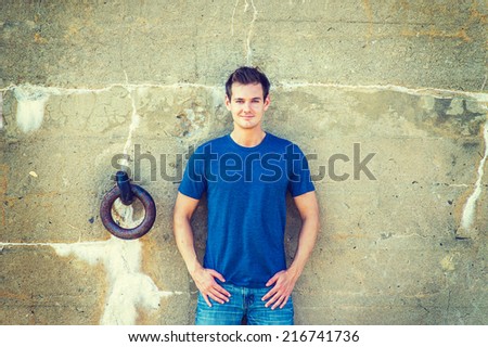 Portrait of Young Sexy Man.  Wearing a blue T shirt, jeans, a young handsome guy is standing against a old concrete wall with a rusty metal ring, charmingly looking at you.