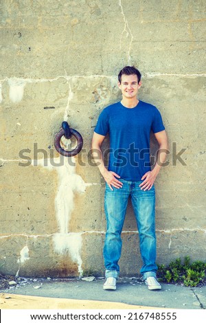Man Casual Fashion. Wearing a blue T shirt, jeans, white sneakers, a young handsome guy is standing against an old concrete wall with a rusty metal ring, charmingly looking at you.