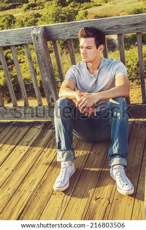 Man Thinking Outside. Wearing a gray T shirt, jeans, white sneakers, a young handsome guy is sitting on the wooden floor,  back against fence in a remote location, relaxing, thinking, lost in thought.