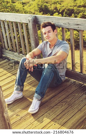 Man Relaxing Outside. Wearing a gray T shirt, jeans, white sneakers, a young handsome guy is sitting on the wooden floor, back against fence in a remote location, relaxing.