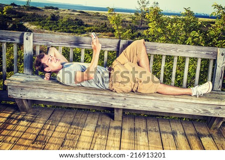 Man Relaxing Outside. Wearing a gray T shirt, yellow pants, white sneakers, a young guy is lying down on wooden bench against fence in remote location, checking messages on mobile phone.