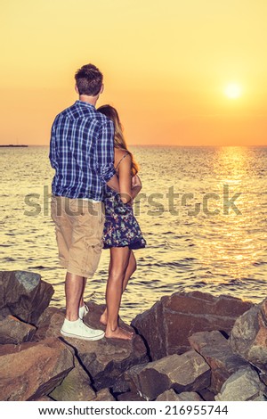 Couple Watching Sunset in Back View. Guy wearing shirt, shorts, sneakers, girl dressing in sundress, barefoot, a couple relying on each other, standing on rocks on beach, looking horizontal of ocean.