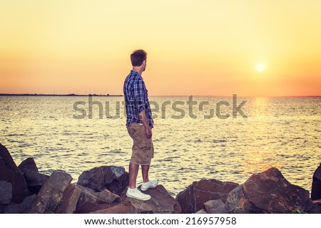 Man Watching Sunset in Back View. Wearing white, blue patterned shirt, yellow pants, white sneakers, a handsome guy is standing on rocks on beach, looking at horizontal of ocean, waiting for you.