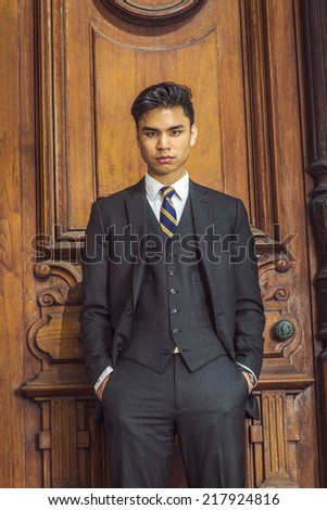 Portrait of Young Business Man. Dressing in a black suit, patterned necktie, vest, hands in pockets, a young handsome businessman is standing by an old fashion style office door, looking at you.