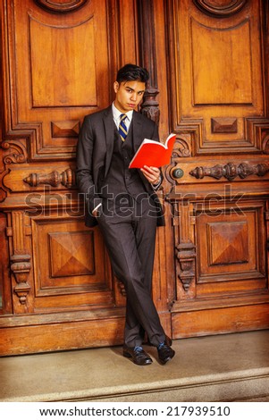 Man Reading. Dressing in a black suit, patterned necktie, vest, leather shoes, a young handsome college student is standing by an old fashion style office door, crossing legs, reading a red book