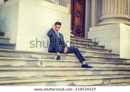 College Student Thinking Outside. Dressing in a black suit, patterned necktie, leather shoes, a young businessman is sitting on stairs outside an office, hand scratching head, relaxing, thinking.