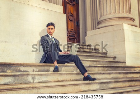 College Student Working Outside. Dressing in a black suit, patterned necktie, leather shoes, a young businessman is sitting on stairs outside an office, working on a laptop computer, thinking.